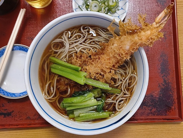 「能登屋支店」の「天ぷらそば」（並）と「キリンラガー」でランチ。大きな「海老天」が一本、潔く鎮座した「天ぷらそば」の登場だ。その「海老」の立派さに「ほう、やるものだな」と独りごち、早速「蕎麦」を手繰り始める。途中で青さの際立った「小松菜」もいただけば、いや、うまいのだよ（笑）。（千束通り商店街：浅草四丁目）